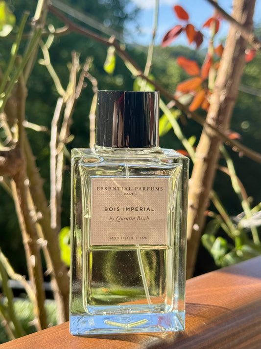 A clear glass perfume bottle with a black cap and a beige label reading 'ESSENTIAL PARFUMS PARIS BOIS IMPERIAL by Quentin Bisch', placed on a wooden surface with blurred greenery and branches in the background.