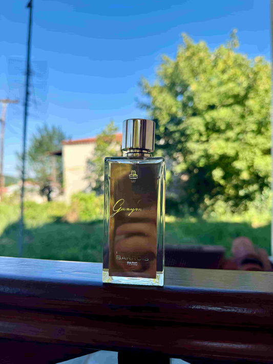 A rectangular glass perfume bottle with a shiny silver cap stands on a dark wooden ledge outdoors. The bottle has a gold-toned label with the brand name Marc-Antoinne Barrois and the scent name Ganymede.